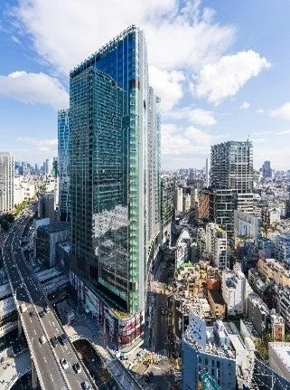 晴れた日の都市全景。ガラス張りの高層ビルがそびえ立ち、車が走る幹線道路が隣接。遠方には密集したビル群が広がり、現代的な大都市の活気ある景観を捉えています。